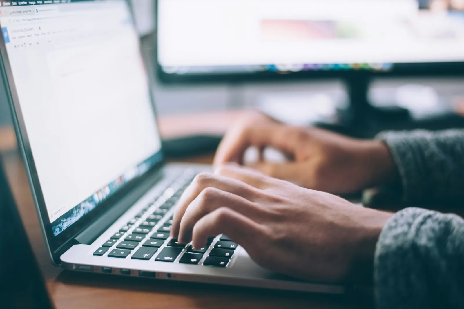 Hands typing on a laptop at a desk, with a second monitor in the background.