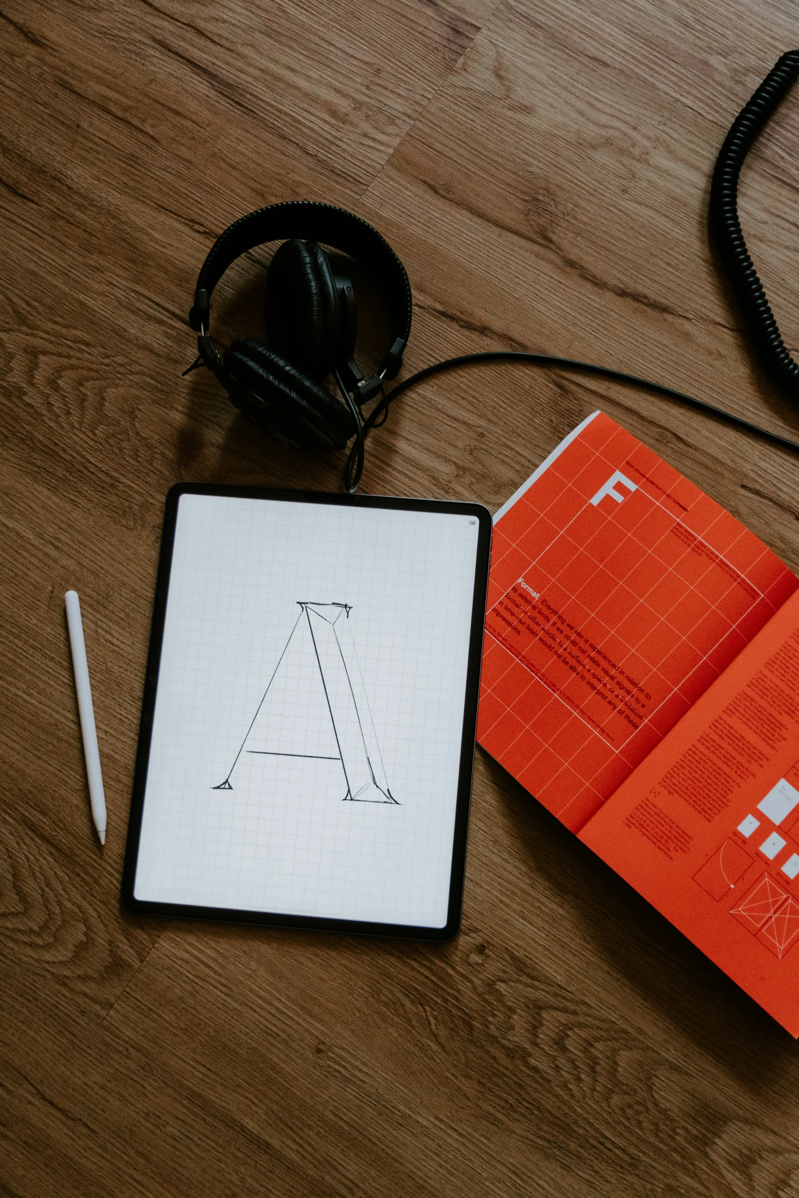 A tablet with a sketched letter A, a stylus, headphones, and an open design book on a wooden desk.